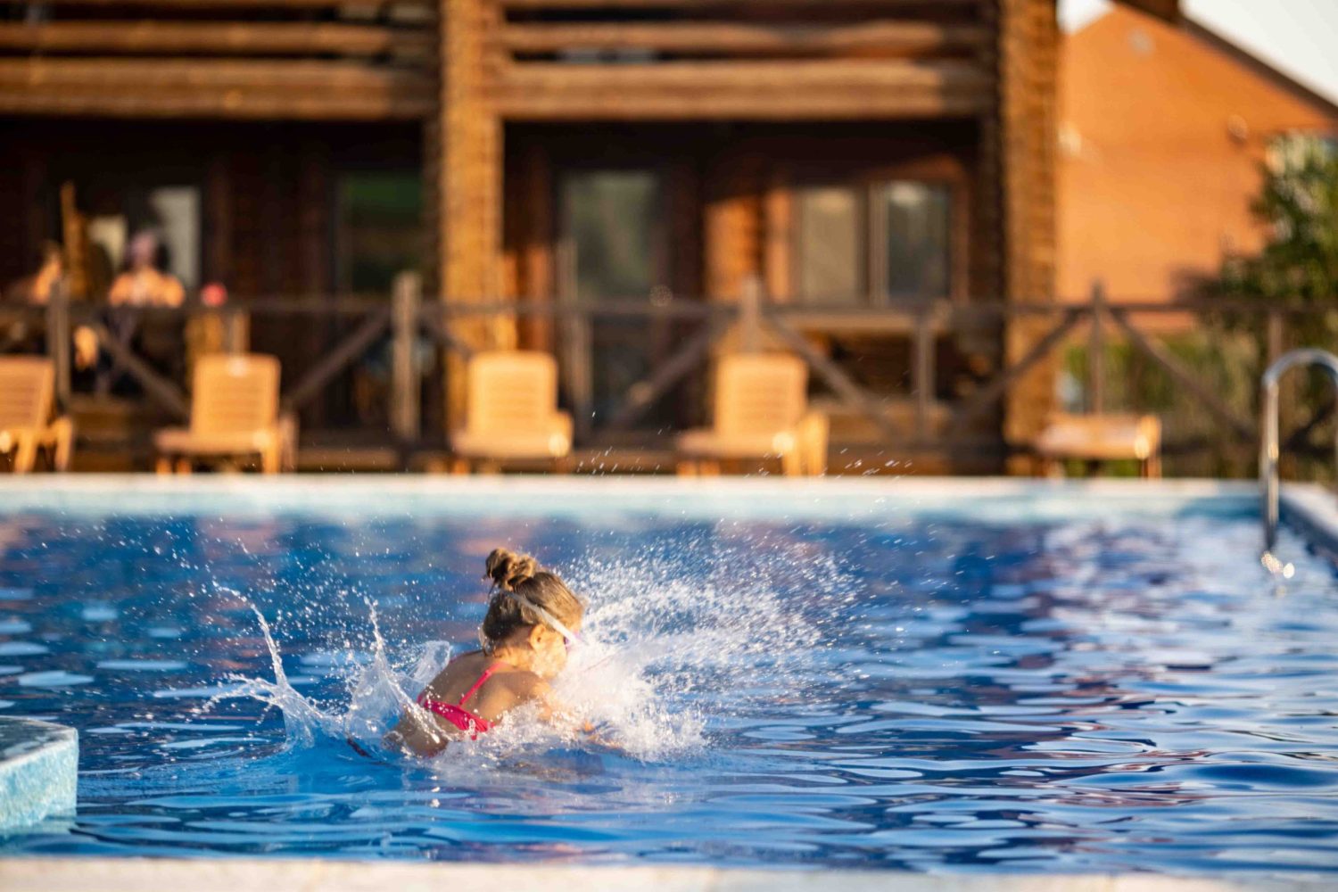 A girl with swimming goggles jumps into a pool with clear water on the background of a warm summer A girl in bright pink swimsuit and black swimming goggles, funny jumping into deep blue pool with clean transparent cool water against background of the warm summer sunny sunset