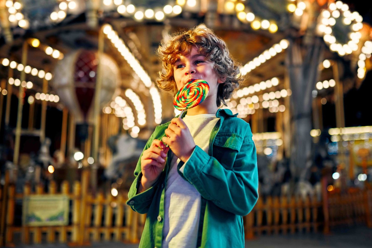 Kids having fun on a carnival Carousel A cute caucasian boy with blonde curly hair eating a colorful lollipop standing against the background of a carousel with horses in the evening at an amusement park or circus.